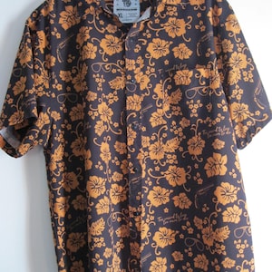 May include: Dark navy button-down shirt with a repeating pattern of orange flowers, eyeglasses, and guns. The shirt has short sleeves and a chest pocket. The text "Too weird to live, too rare to die" is printed on the shirt.
