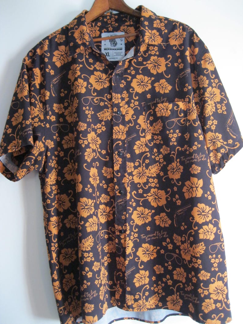 May include: Dark navy button-down shirt with a repeating pattern of orange flowers, eyeglasses, and guns. The shirt has short sleeves and a chest pocket. The text "Too weird to live, too rare to die" is printed on the shirt.