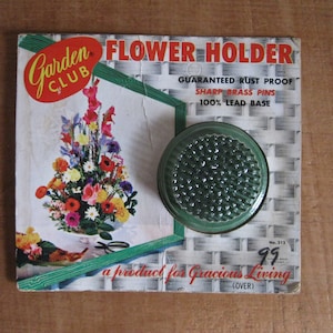 Op de afbeelding: Vintage bloemenhouder met een groene glazen kikker. De verpakking toont een bloemstukillustratie en de tekst: "GEGARANDEERD ROESTVRIJ, SCHERPE MESSING PENNEN, 100% LOOD BASIS". De verpakking leest "Garden Club".