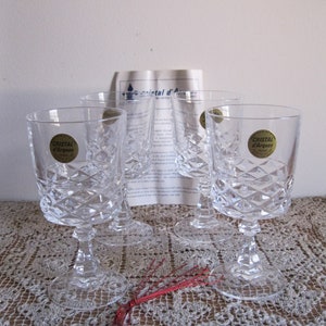 May include: Set of four clear crystal glasses with a diamond pattern. Each glass has a gold label that reads "Cristal d'Arques". The glasses are on a white lace tablecloth.