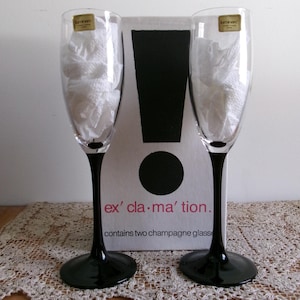 May include: Two champagne flutes with black stems and bases, filled with white paper, stand before a box with a black exclamation point design. The box reads "ex' cla-ma' tion" and "contains two champagne glasses."