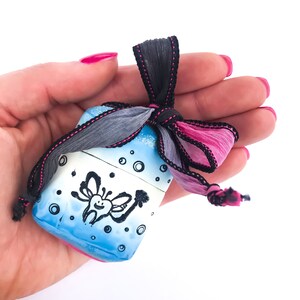 Op de afbeelding: Een klein blauw en wit geschenkdoosje met een zwart-witte tekening van een lachende tand met vleugels. Het doosje is vastgebonden met een zwart lint met roze rand.