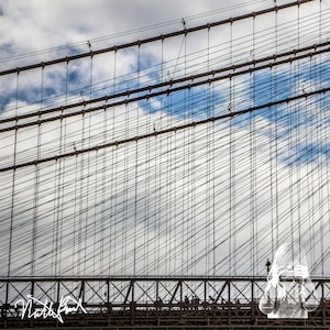 May include: A black and white photo of a suspension bridge with a complex network of cables and beams. The bridge is against a cloudy sky.