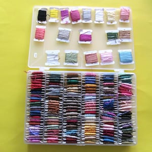May include: A clear plastic storage box filled with spools of colourful embroidery floss. The box has two levels, with the top level containing 16 spools and the bottom level containing 48 spools.
