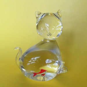 May include: A clear glass cat figurine with a red and white fish inside. The cat is sitting with its tail curled up. The figurine is on a yellow background.