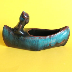 May include: A green and brown ceramic canoe planter with a small figurine of a person paddling. The canoe is inscribed with "Merrill, WI".