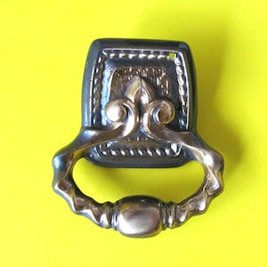 May include: A black metal drawer pull with a decorative gold fleur-de-lis design and a ring handle.