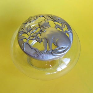 May include: A clear glass jar with a pewter lid featuring a cat design. The cat is sitting on a branch with leaves and is looking up at a butterfly.