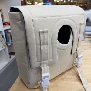 Può includere: Uno zaino in tela beige con un'apertura circolare e fibbie in plastica bianca. Lo zaino ha una forma rettangolare con una parte superiore curva e una fodera interna blu scuro. Le cinghie sono regolabili.