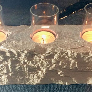 Puede incluir: Un portavelas de piedra con tres portavelas de vidrio. El portavelas está hecho de piedra rugosa y texturizada y tiene una forma rectangular. Los portavelas de vidrio son transparentes y tienen una forma redonda. Las velas están encendidas y emiten un brillo cálido.