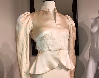May include: A cream-colored satin blouse with a V-neck and long sleeves. The sleeves have a puffed shoulder and are embellished with small, white sequins.