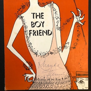 May include: A vintage program for the musical "The Boy Friend" featuring a stylized illustration of a woman in a pink dress with a black outline and a signature by the actress Phylis Newman. The background is orange.