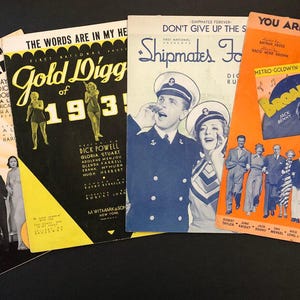 May include: Four vintage sheet music covers with colorful illustrations and text. The covers feature musical titles like "Wonder Bar", "Gold Diggers of 1933", "Shipmates Forever", and "Broadway Melody of 1936".