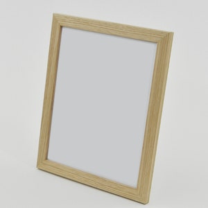 May include: A rectangular wooden picture frame with a light-coloured, natural wood grain finish. The frame has a simple, clean design and is angled slightly, showcasing the white interior space for a photograph or artwork. Suitable for home decor.