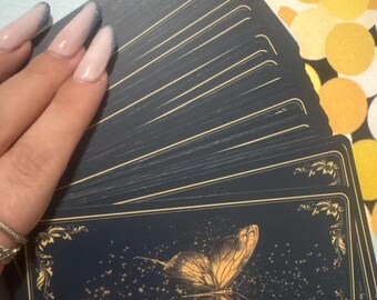 May include: A deck of tarot cards with a navy blue background and gold accents. The cards feature a gold butterfly design on the front. The deck is fanned out, with a hand gently touching the cards. The nails are painted with a light pink and blue glitter.