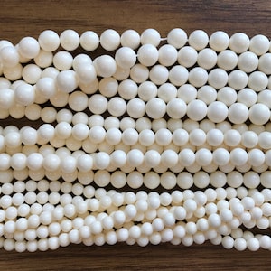 May include: A close-up of a strand of white round beads. The beads are smooth and have a slight sheen. The beads are arranged in a random pattern.