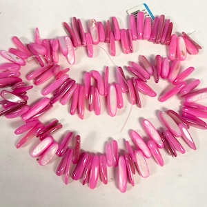 May include: A strand of pink shell beads. The beads are elongated and vary in shades of pink, with some featuring a pearlescent white center. The beads are strung together on a thin, clear string, creating a necklace.