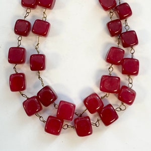 May include: A two-strand necklace featuring square, burgundy-colored beads. The beads are strung on a delicate, gold-toned chain. The necklace is displayed against a white background, highlighting the rich color and design of the jewelry.
