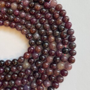 May include: A strand of round, purple gemstone beads. The beads are smooth and have a slight sheen. They are arranged in a circle, with the ends of the strand overlapping.