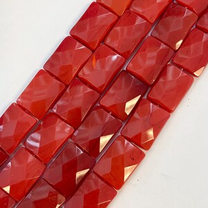May include: Close-up of a collection of faceted, rectangular red beads. The beads have a glossy finish and are arranged in rows, showcasing their geometric cut. Ideal for jewelry making, these beads offer a vibrant color and unique texture.