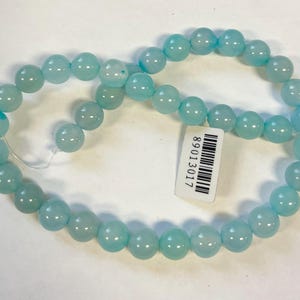 Op de afbeelding: Een streng lichtblauwe ronde kralen van agaat. De kralen zijn geregen op een wit draad. De kralen zijn ongeveer 10 mm in diameter. De streng is ongeveer 45 cm lang. De kralen zijn glad en hebben een lichte glans. De streng is perfect voor het maken van sieraden of andere knutselwerkjes.