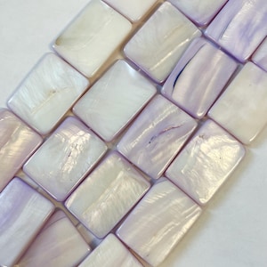May include: A collection of rectangular mother-of-pearl beads arranged in a grid pattern. Each bead features a smooth, iridescent surface with a lustrous finish, displaying soft shades of white and light purple with natural variations in texture.