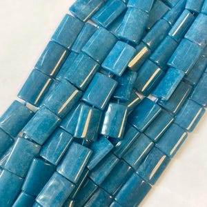 May include: A close-up of a collection of rectangular, blue-green gemstone beads. The beads are strung together, showing their polished, smooth surfaces. The colour is a vibrant, sea-like hue, ideal for jewellery making.
