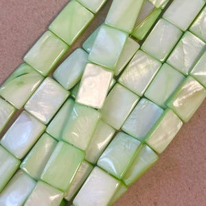 May include: Close-up of a collection of rectangular, iridescent beads in shades of pale green and white. The beads have a glossy finish and are arranged in a diagonal pattern, suggesting they are strung together for jewelry making.