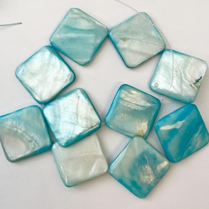 May include: A collection of square, iridescent beads in shades of light blue and white. The beads are strung together, showcasing their glossy, polished surfaces. The beads have rounded corners and a pearlescent sheen.