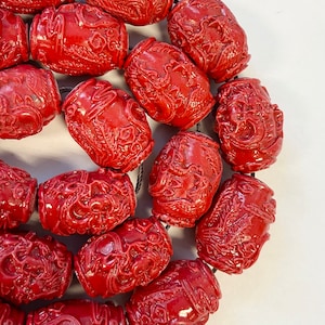 以下が含まれることがあります： 複雑なデザインが施された赤い円筒形のビーズのネックレス。ビーズは黒い紐で繋がっています。