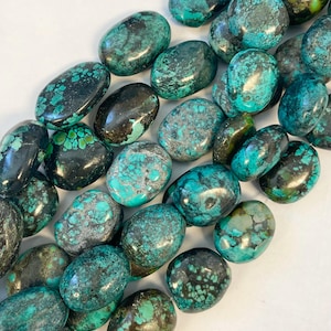 May include: A collection of polished turquoise gemstones with a mix of dark and light blue-green hues. The stones have a smooth, oval shape and varying patterns, with some displaying dark matrix veining. The gemstones are arranged closely together, filling the frame.