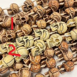May include: A string of beige and brown skull beads with intricate carvings. The beads are strung on a white cord and are approximately 1 inch in diameter.
