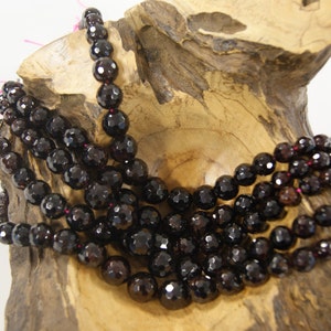 May include: A strand of dark red gemstone beads, likely garnet, displayed on a wooden surface. The beads are round and faceted, creating a shimmering effect.