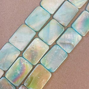 May include: Close-up of rectangular iridescent beads with a pearlescent sheen. The beads have a light blue hue with hints of yellow and green, and are arranged in rows. These beads are ideal for jewelry making.
