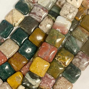 May include: A collection of square-cut gemstones in various colors, including green, yellow, red, and brown. The stones have a polished, glossy finish and are arranged closely together. The gemstones are likely for jewelry making or crafting.