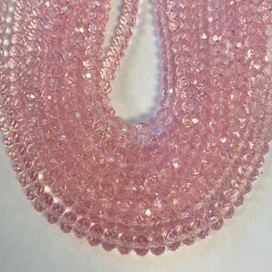 May include: Close-up of multiple strands of light pink faceted beads. The beads are strung together, creating a layered effect. The beads have a sparkling, iridescent quality, reflecting light. The overall effect is delicate and feminine.