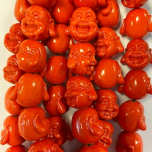 Puede incluir: Una hebra de cuentas redondas de color naranja con un diseño de cara de Buda sonriente. Las cuentas están enhebradas en un cordón negro.