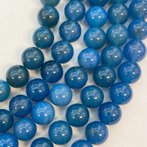 May include: A collection of glossy, round beads in various shades of blue, from deep teal to light sky blue. The beads are arranged closely together, reflecting light and creating a smooth, polished appearance. Ideal for jewelry making or crafting.