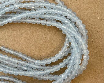 Pode incluir: Um fio de contas de pedra preciosa facetadas de cor azul claro. As contas são redondas e têm um acabamento liso e polido.