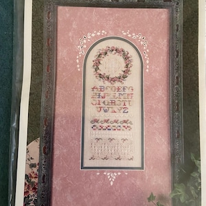 May include: A cross-stitch pattern titled Memories of Spring by Amie Bishop, featuring an arched design with a floral wreath, the alphabet, and decorative borders. The pattern is displayed in a dark frame against a pink marbled background.