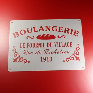 Op de afbeelding: Witte sjabloon met de tekst "BOULANGERIE", "LE FOURNIL DU VILLAGE", "Rue de Richelieu" en "1913". De sjabloon bevat ook een gestileerd brood en decoratieve versieringen.