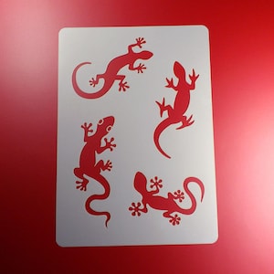 Könnte beinhalten: Weiße Schablone mit vier Silhouetten von Eidechsen. Die Eidechsen sind in verschiedene Richtungen ausgerichtet und haben unterschiedliche Posen. Die Schablone besteht aus einem dünnen, flexiblen Material und ist für die Verwendung mit Farbe oder anderen Medien konzipiert.