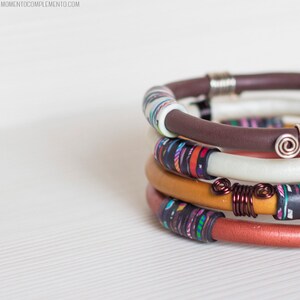 以下が含まれることがあります： 茶色、白、オレンジ、赤のバンドが付いた、カラフルなブレスレットが5つ重ねられています。各ブレスレットは、カラフルなビーズのユニークなパターンで、銀と銅のワイヤーでアクセントが付けられています。