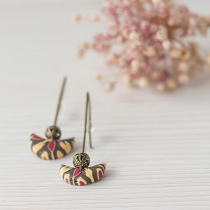 May include: A pair of dangle earrings with a crescent moon shape. The earrings are made of polymer clay and have a beige, red, and black pattern. The earrings have a gold-colored metal hook.