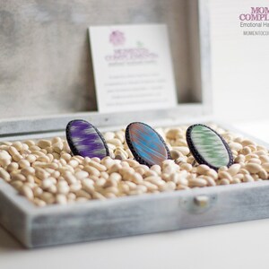 May include: Three oval-shaped handmade jewelry pieces with colorful designs, displayed in a gray wooden box filled with white beans. The jewelry features purple, blue, and green patterns with black borders. The box also contains a small card with text.