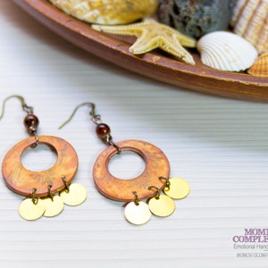 Könnte beinhalten: Handgefertigte Ohrringe mit orangefarbenen und braunen kreisförmigen Anhängern mit floralem Design und kleinen goldfarbenen Scheiben. Die Ohrringe haben bronzefarbene Haken und eine kleine braune Perle.