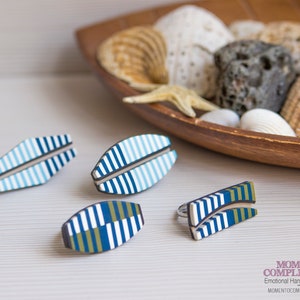 May include: Four pieces of handmade jewelry with a nautical theme. The pieces include a ring and three other decorative pieces. Each piece has a white, blue, and green striped design. A wooden bowl filled with seashells is in the background.
