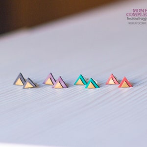 Könnte beinhalten: Handgefertigte Dreiecks-Ohrstecker in verschiedenen Farben, darunter Schwarz, Lila, Türkis und Koralle, jeweils mit goldfarbener Basis. Die Ohrringe sind auf einer weißen Oberfläche präsentiert. Der Text "MOMENTO COMPLEMENTO" ist oben rechts sichtbar.