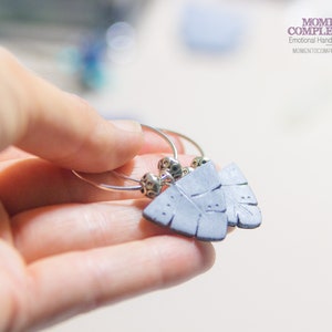 May include: A pair of silver hoop earrings with gray feather-shaped pendants. Each earring features small, decorative silver beads. The earrings are held in a hand, with the brand name "Momento Complemento" visible in the background.