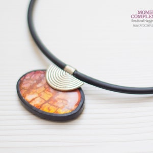 Könnte beinhalten: Eine schwarze Choker-Kette mit einem Anhänger aus orangefarbenem und braunem Harz mit einem silbernen Metallakzent. Der Anhänger hat die Form eines Halbkreises.